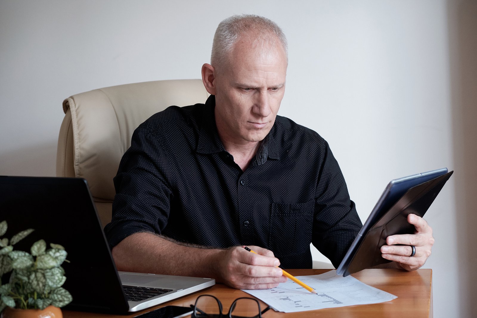 Concentrated mature Caucasian entrepreneur using digital table while sitting at office table and checking data on documents
